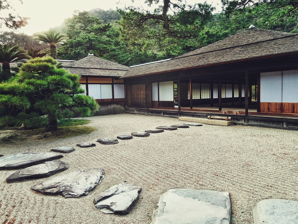 Picture of older house in Japan, large area of gravel stones in front of the house, path made of stone leading to the house's entry. Bonsai tree left to the stone path. Trees or forest in the background, behind the house.